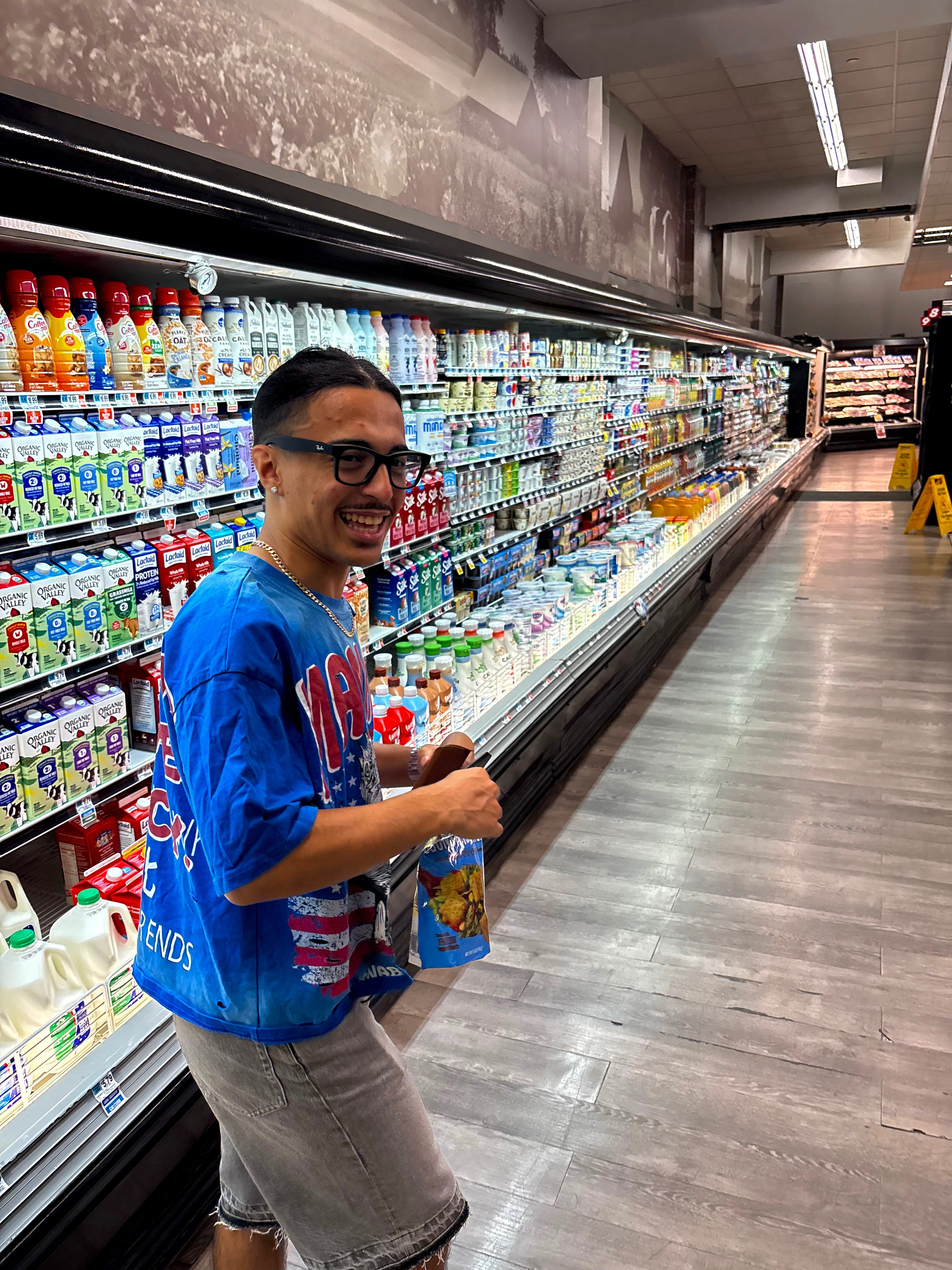 A man wearing a blue graphic t-shirt and gray shorts stands in a grocery store aisle next to a refrigerated beverage display, holding a bottle of juice and a bag of chips.